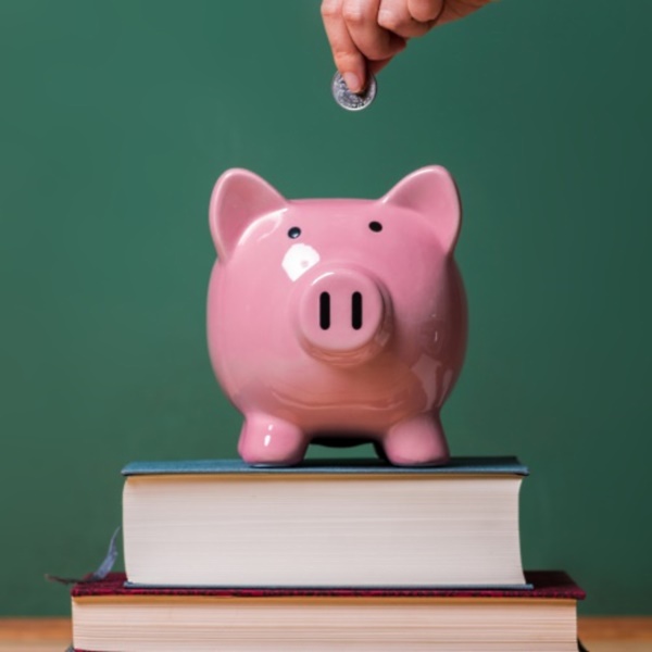 Person depositing money in a pink piggy bank on top of books with chalkboard in the background as concept image of the costs of education